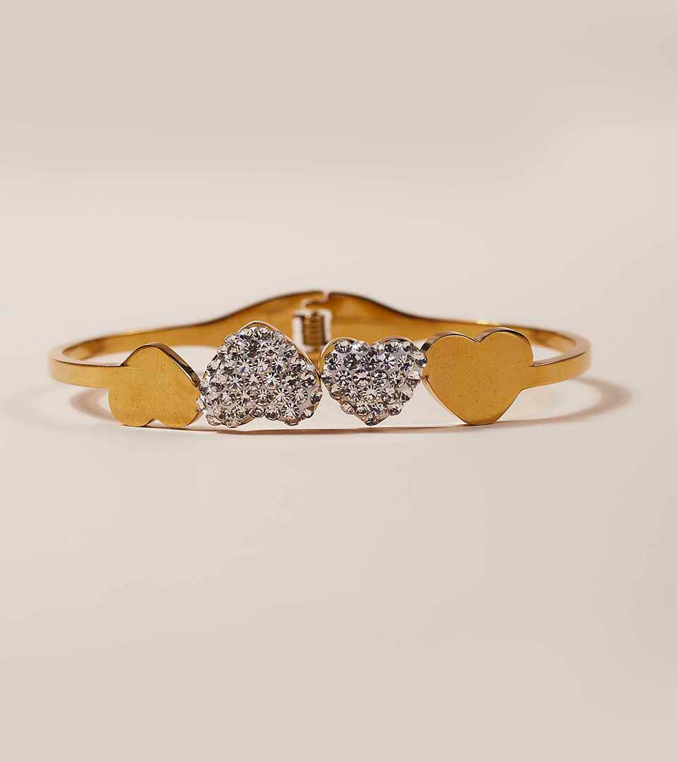 Gold heart-shaped bangle bracelet with two solid gold hearts and two pavé-set clear heart stones in the center, shown on a warm beige background.